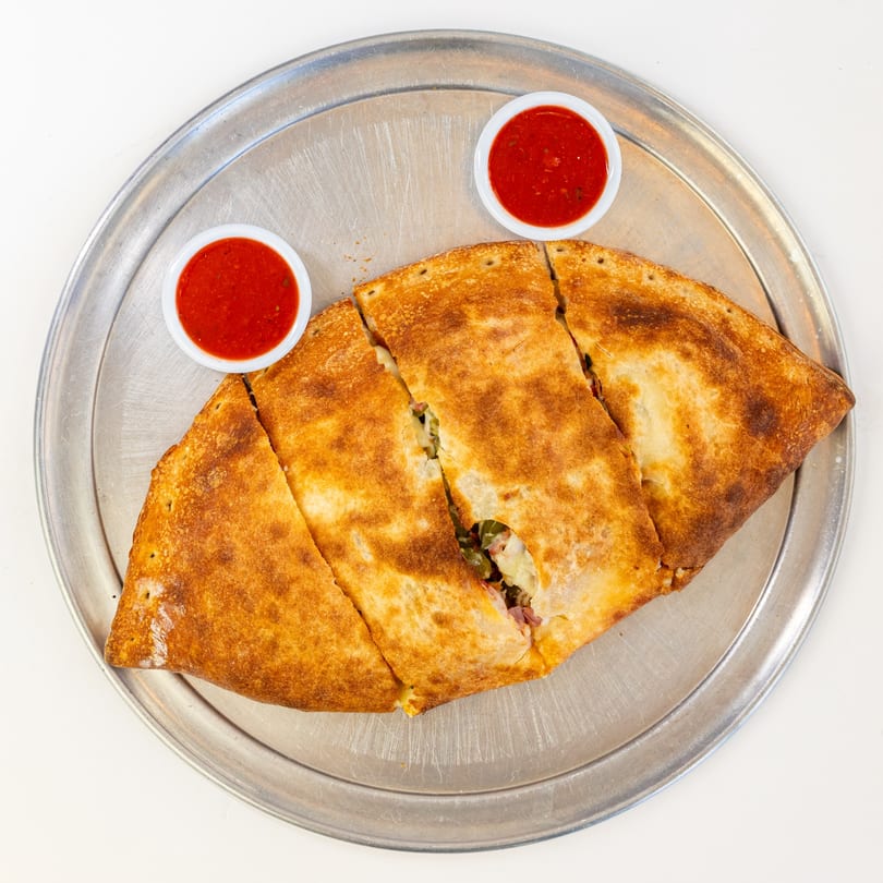A golden-brown calzone, sliced into four pieces, served with two small cups of marinara sauce on a metal pizza pan.