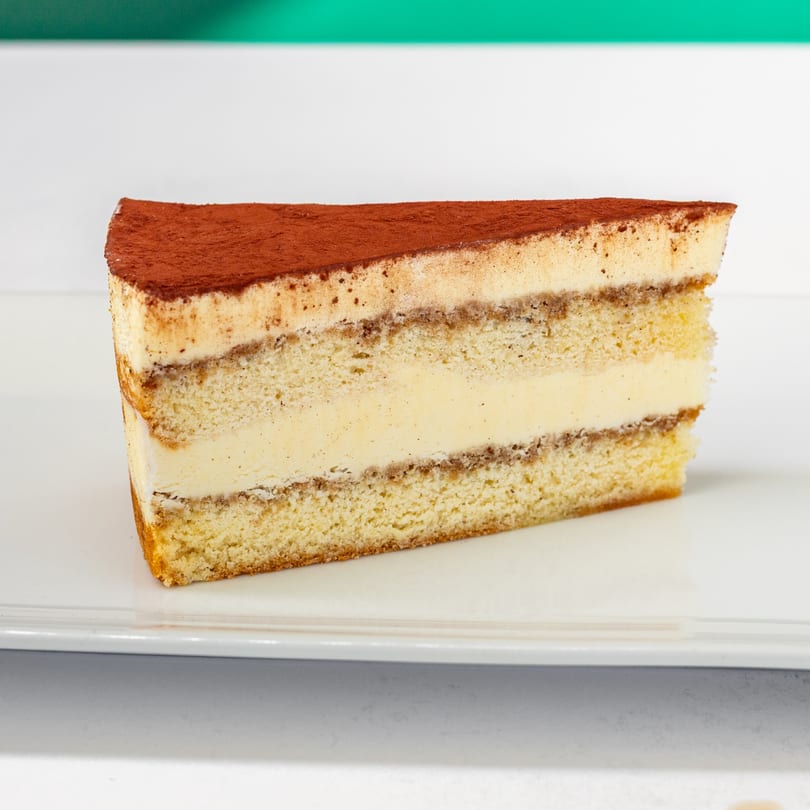 A slice of tiramisu cake with layers of sponge cake, cream, and cocoa powder on a white plate.